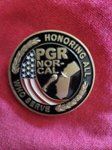 Image of back of Patriot Guard Honor Coin. This coin is a round design, It has wording on the outer edge of circle that reads-Honoring All Who Serve. There are two branches between the words. In the middle section reads-PGR NOR-CAL. The images with the words are-State of CA, with star on it near coastline, in curve of circle is US Flag and between flag and state is a boot with a gun in the boot and a helmet sitting on top of the gun. All lettering and images are in gold.